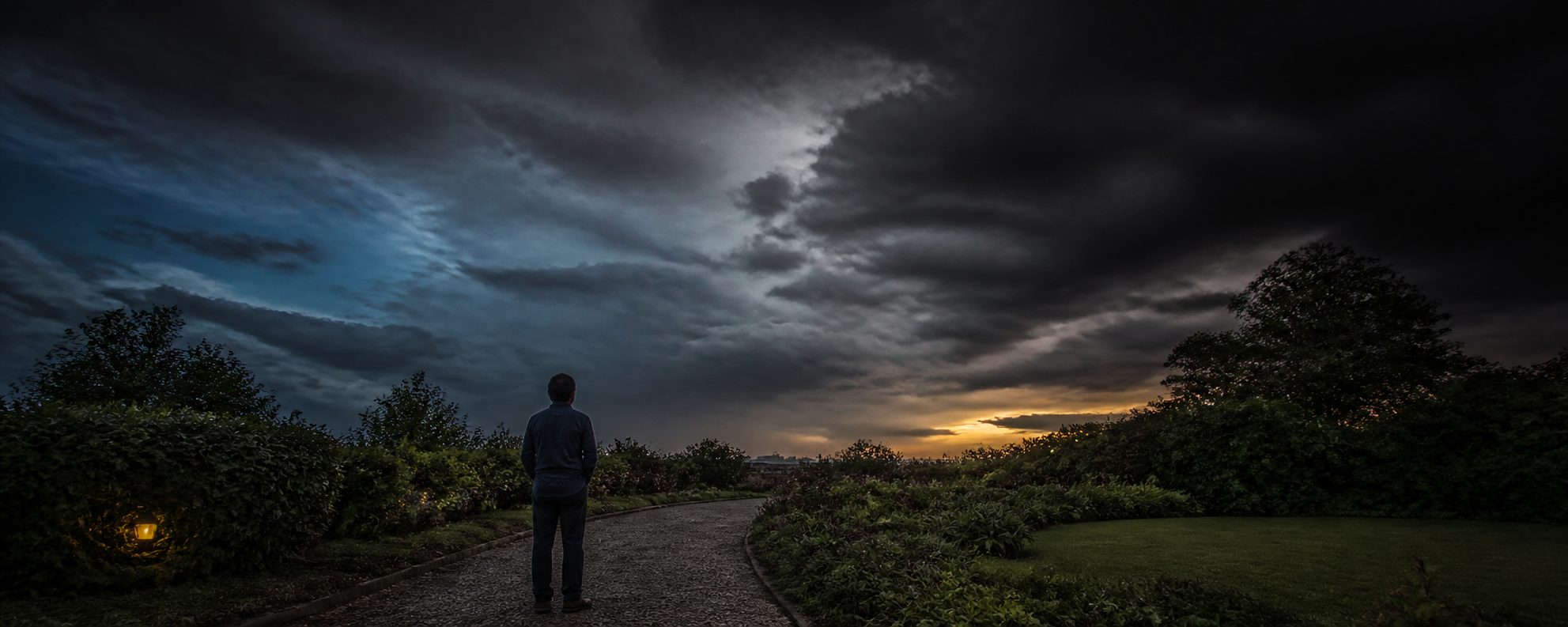 A figure standing alone on a garden path at dusk, facing a vast gray sky of heavy cloud, with a thin band of yellow light still burning along the horizon.